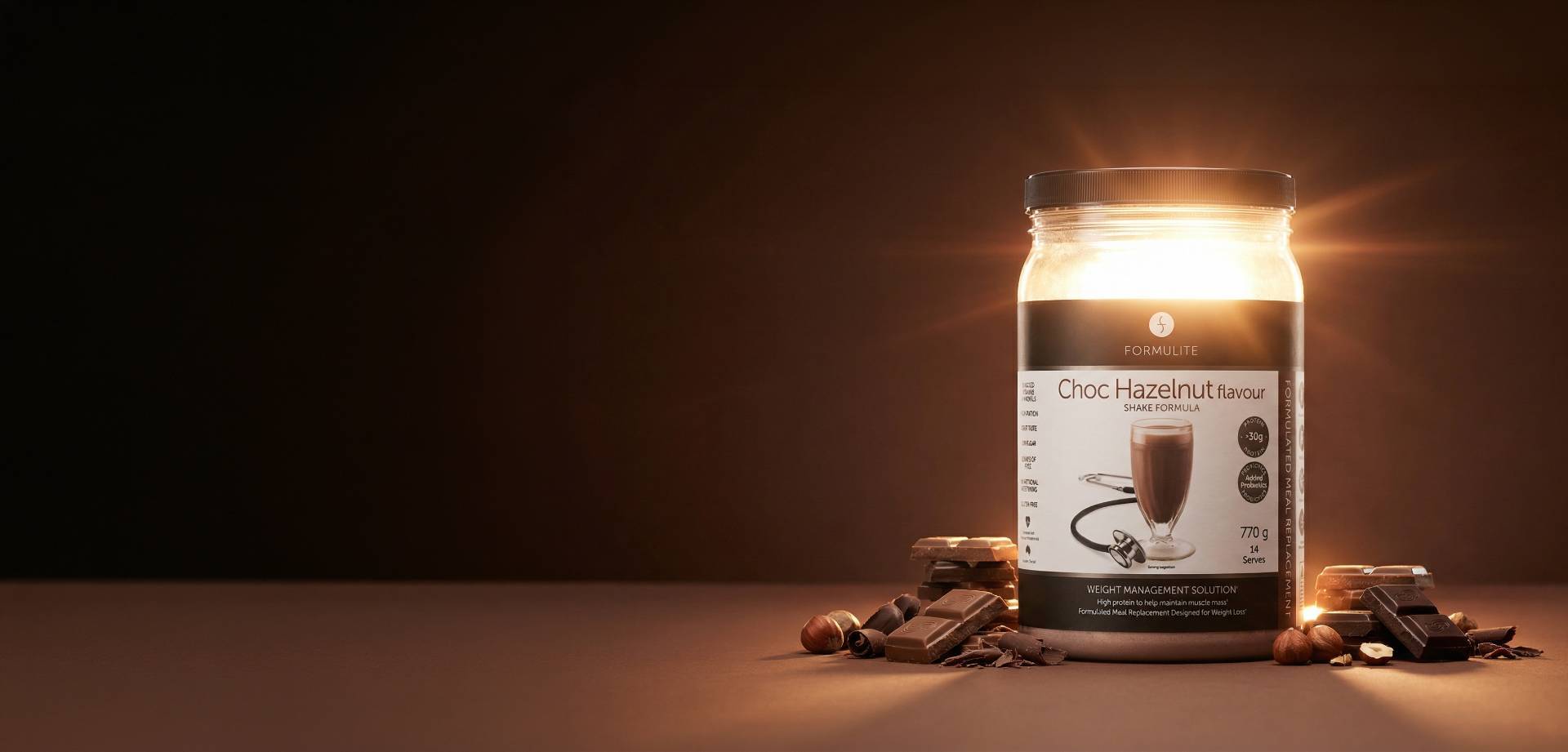 A jar of "Formulite Choc Hazelnut" shake mix with chocolate chunks and hazelnuts. The background is brown.