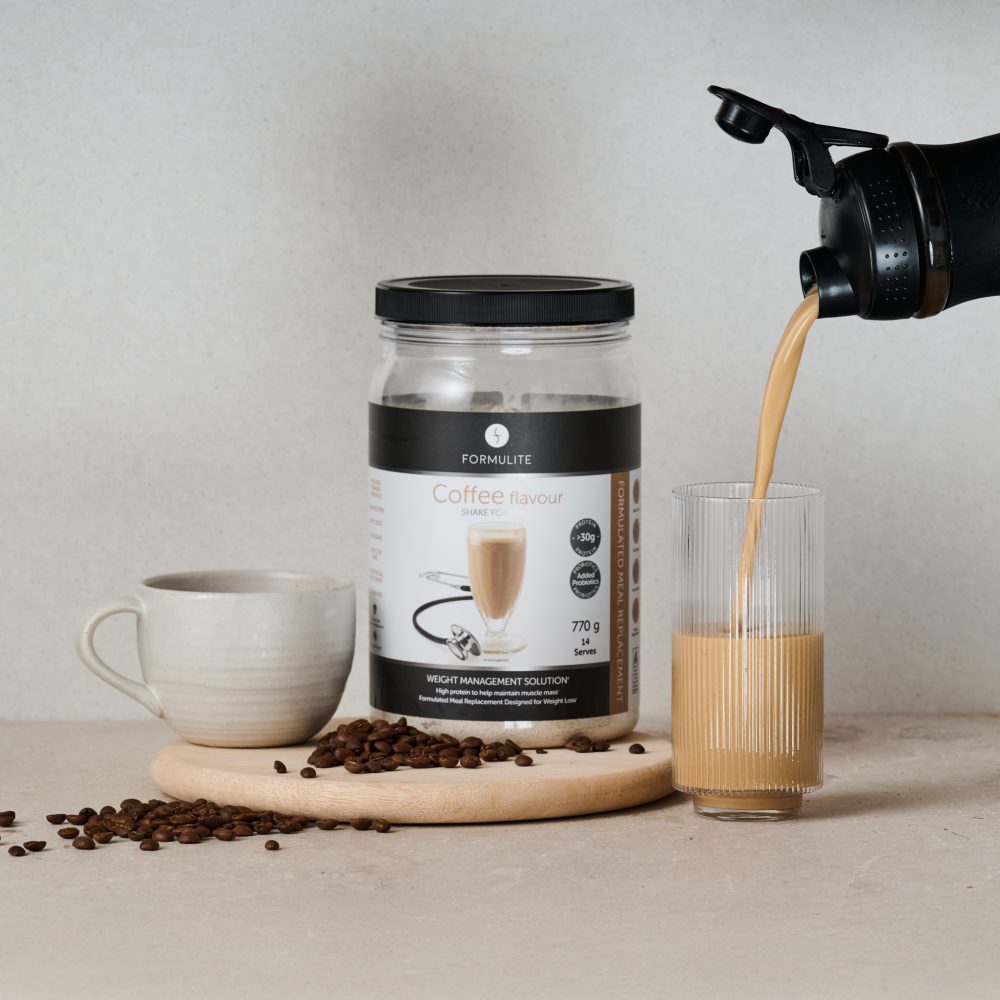 Coffee flavored protein shake being poured into a glass, next to a jar, a cup and coffee beans.