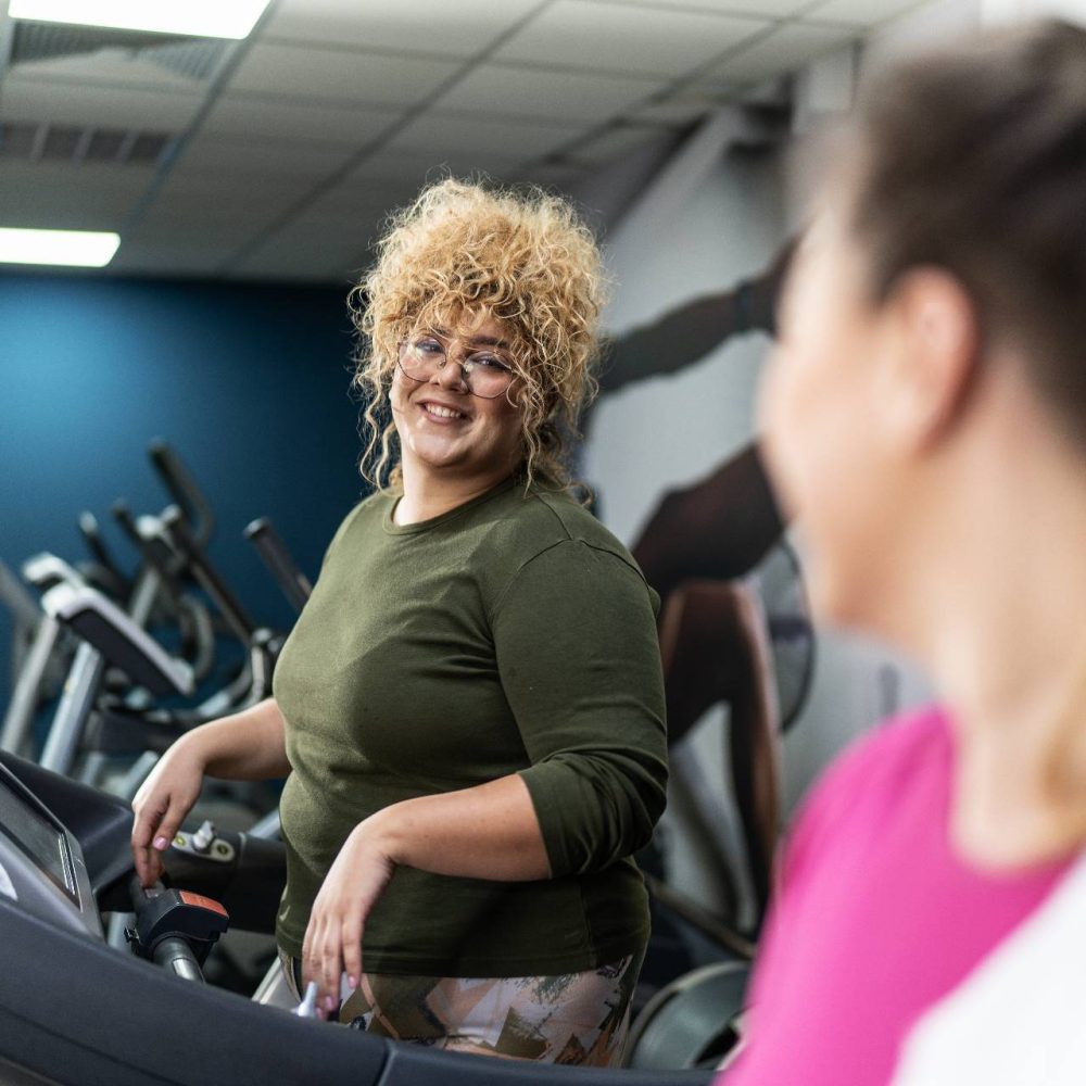 Woman smiles from a treadmill in a gym. Another woman is nearby.